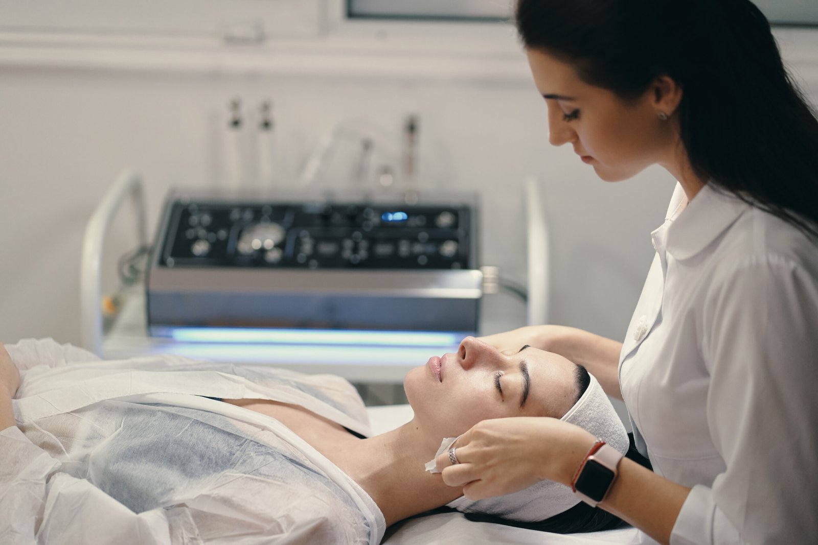 Female cosmetologist making facial treatment to a beautiful woma Woman lying during cosmetic procedure while skillful female cosmetologist making facial treatment